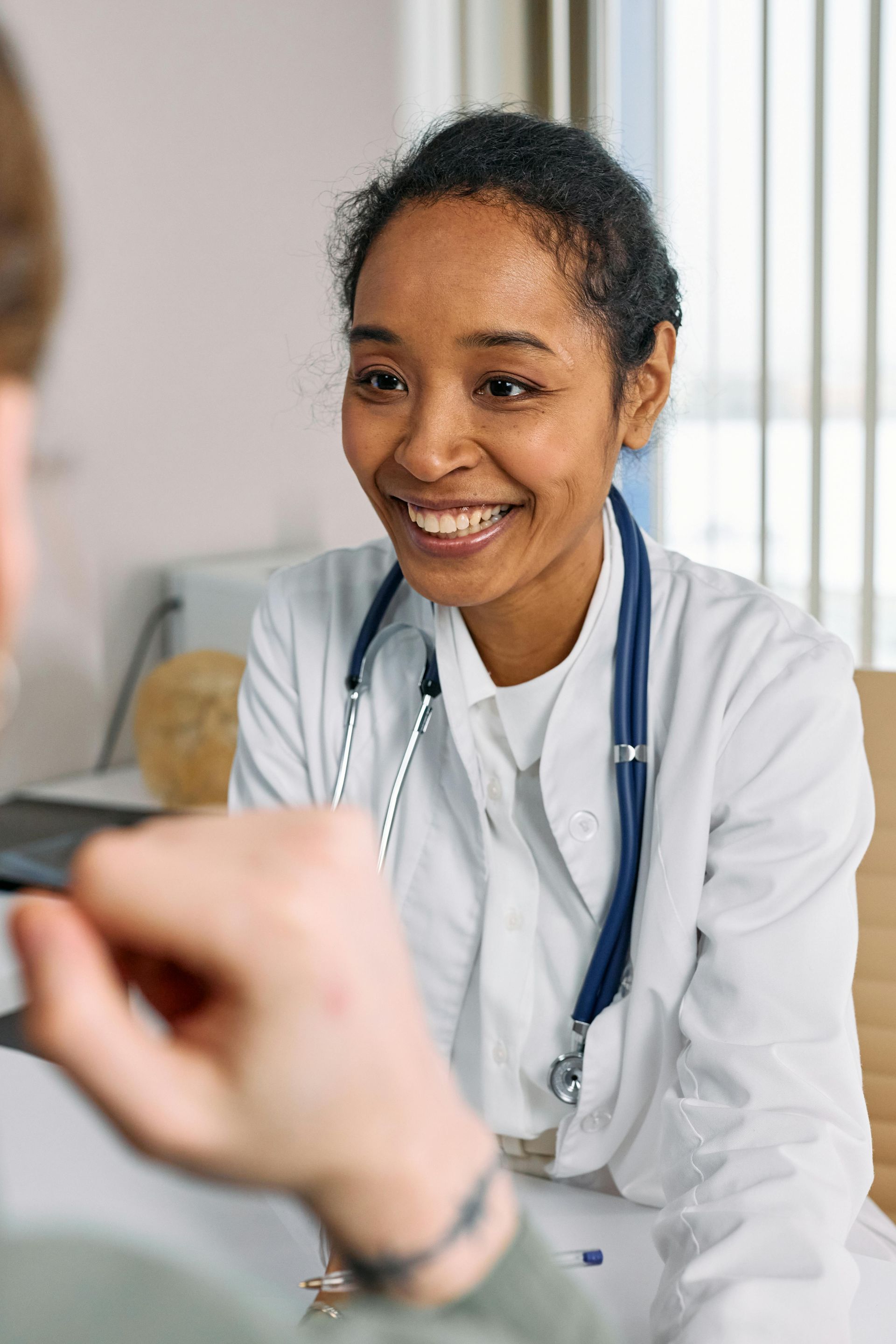 A smiling healthcare professional wearing a white coat and stethoscope speaks with a patient in an office setting.