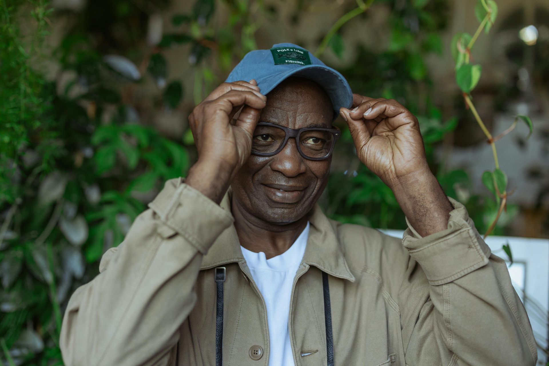 An individual wearing glasses and a tan jacket adjusts a blue cap, set against a backdrop of lush green plants.