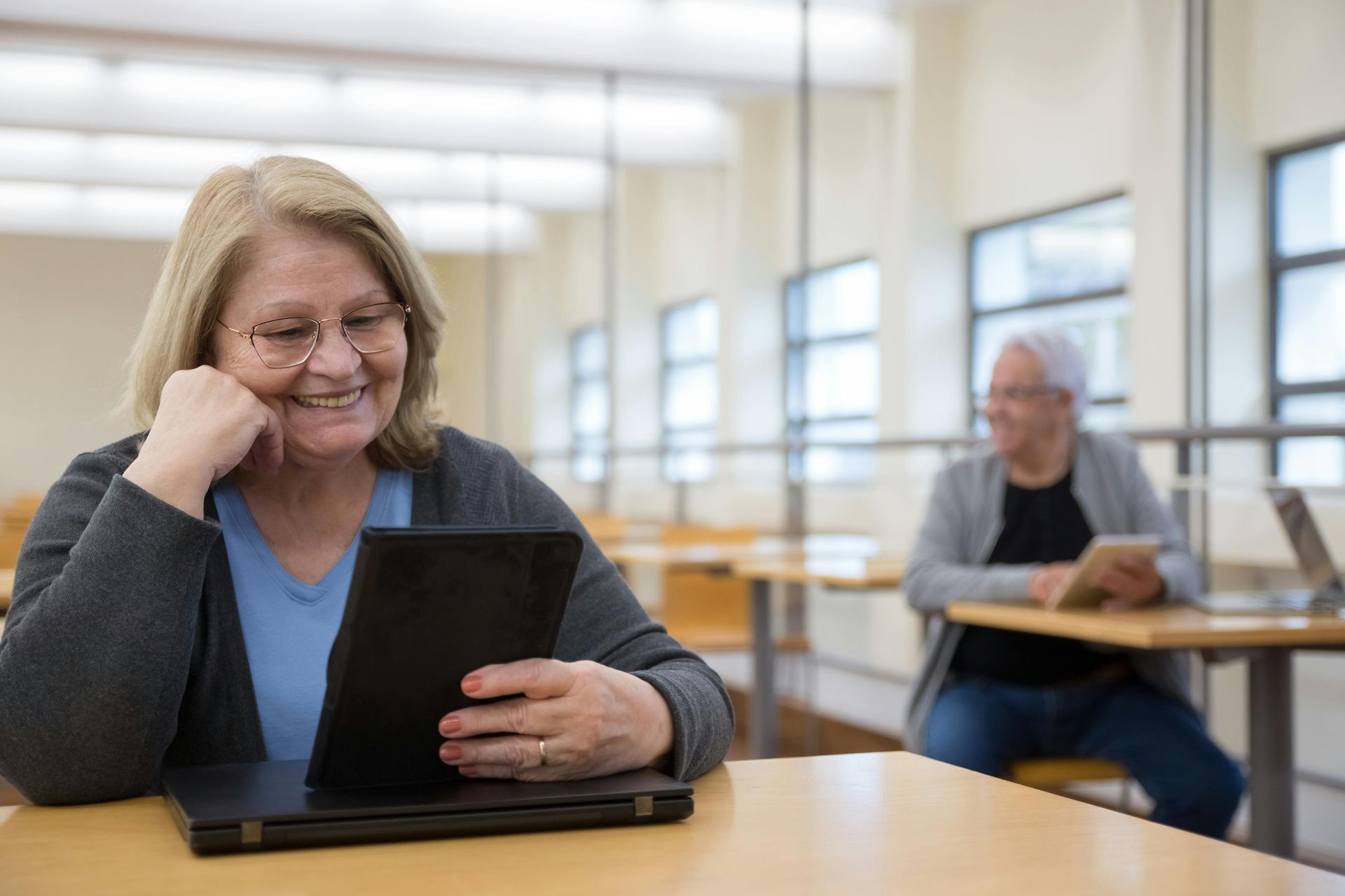 A person sits in a bright library, smiling while reading a digital tablet. Another person sits at a desk in the background.