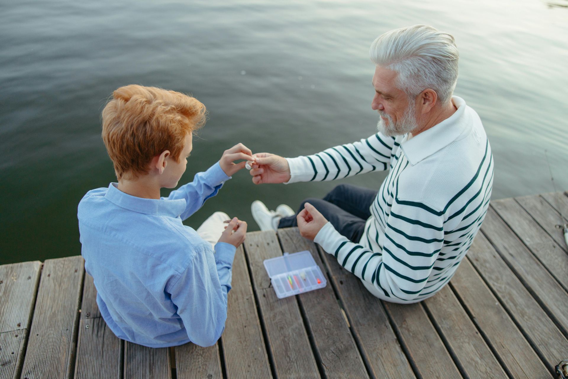 An older adult and a youth sit on a wooden pier by the water, carefully handling fishing equipment together.