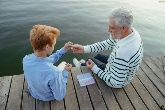 An older adult and a youth sit on a wooden pier by the water, carefully handling fishing equipment together.