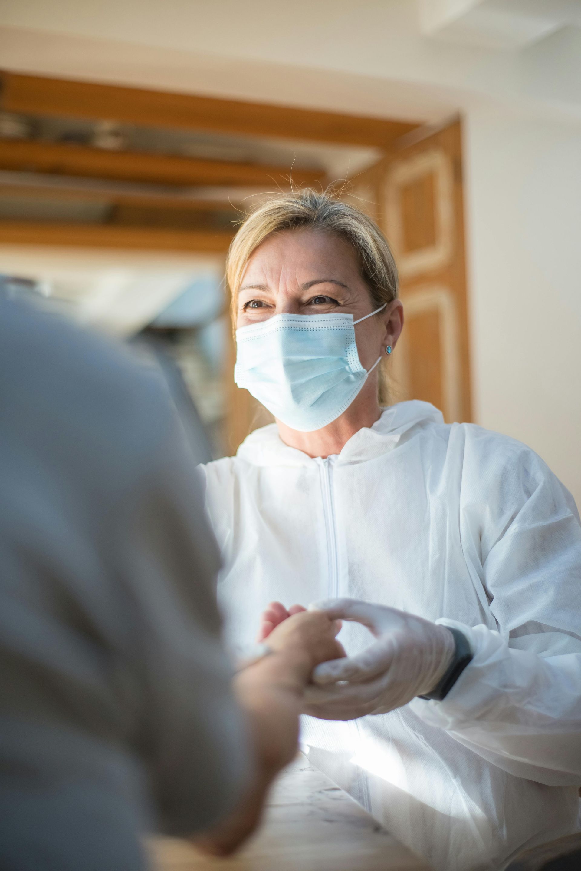 A healthcare worker in a white protective suit and mask gently holds the hand of a person in a bright, indoor setting.