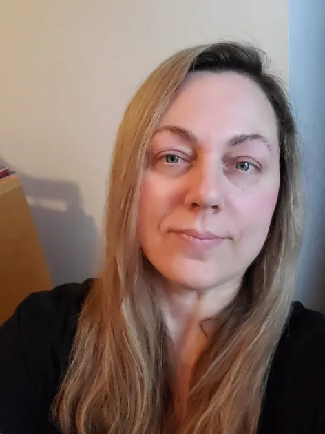 Woman with long blonde hair looking at the camera, neutral expression. Wearing a black top, indoors.