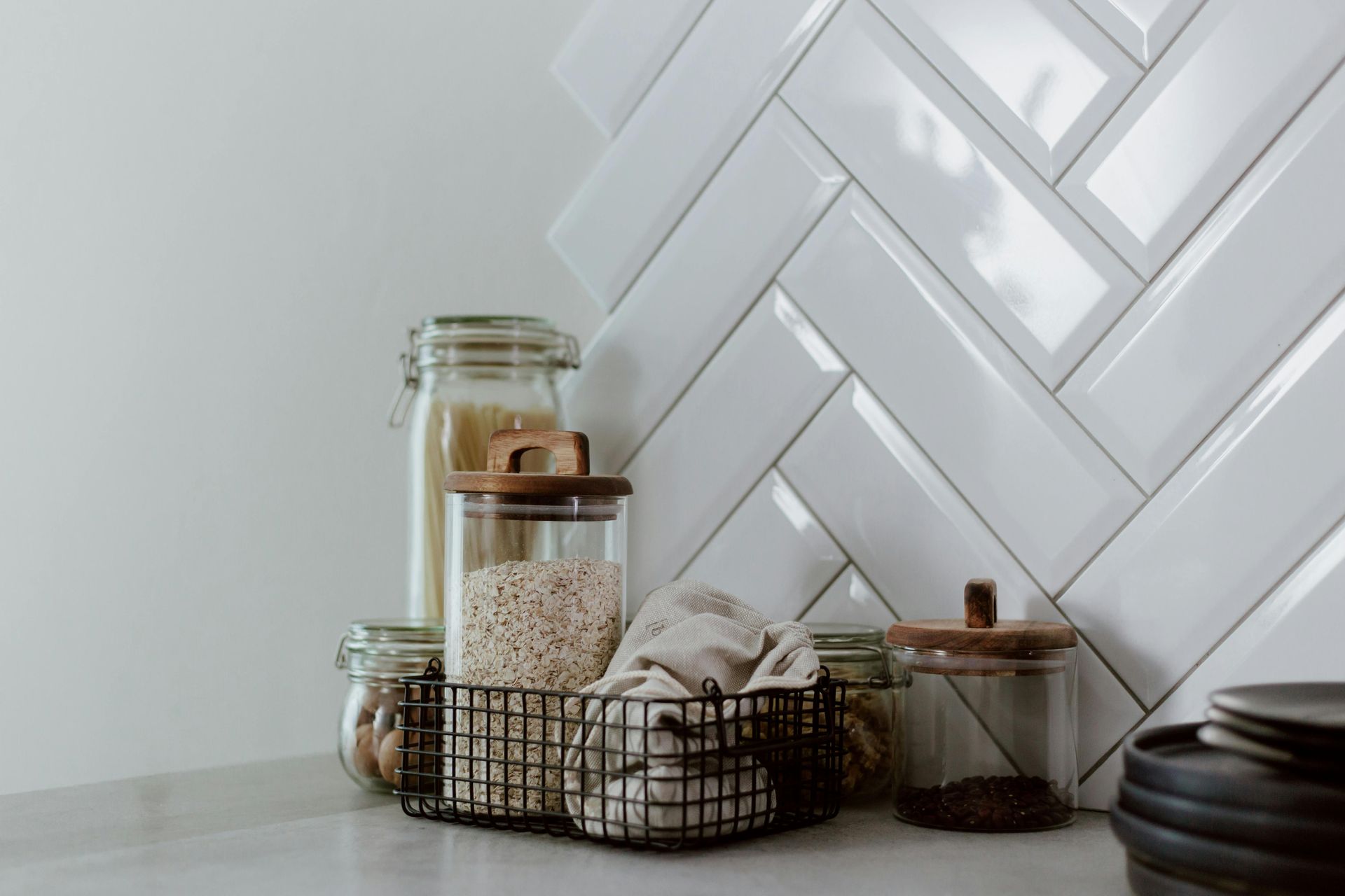 Kitchen counter with glass jars, wire basket, and herringbone tile backsplash.