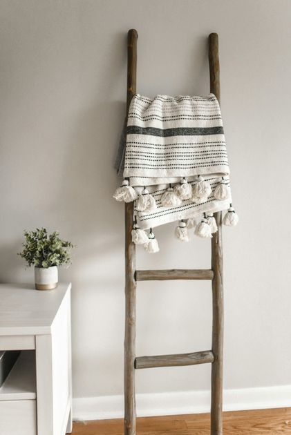 Wooden ladder propped against a gray wall, holding a blanket with tassels; small plant in a white pot.