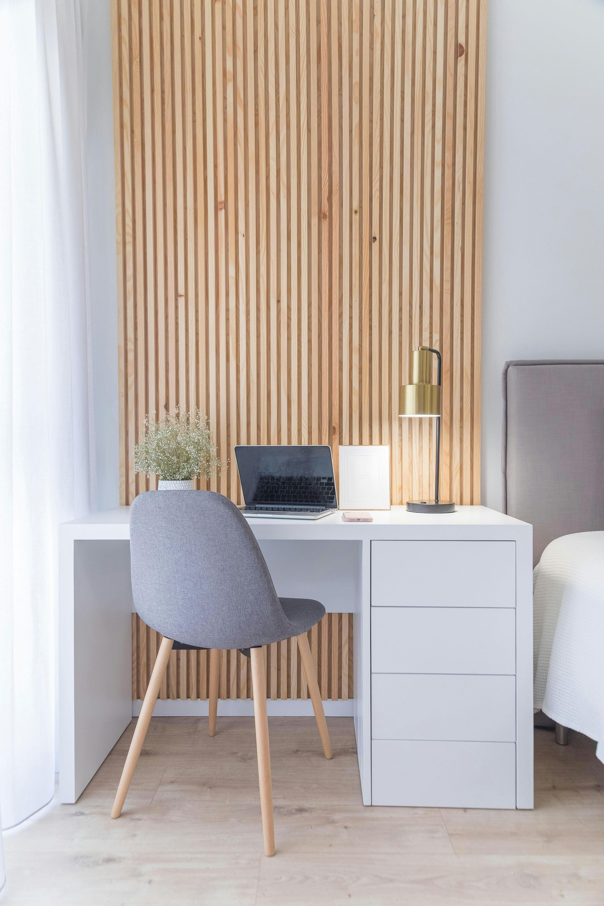 White desk with laptop, gray chair, and gold lamp in front of a wooden slat wall.