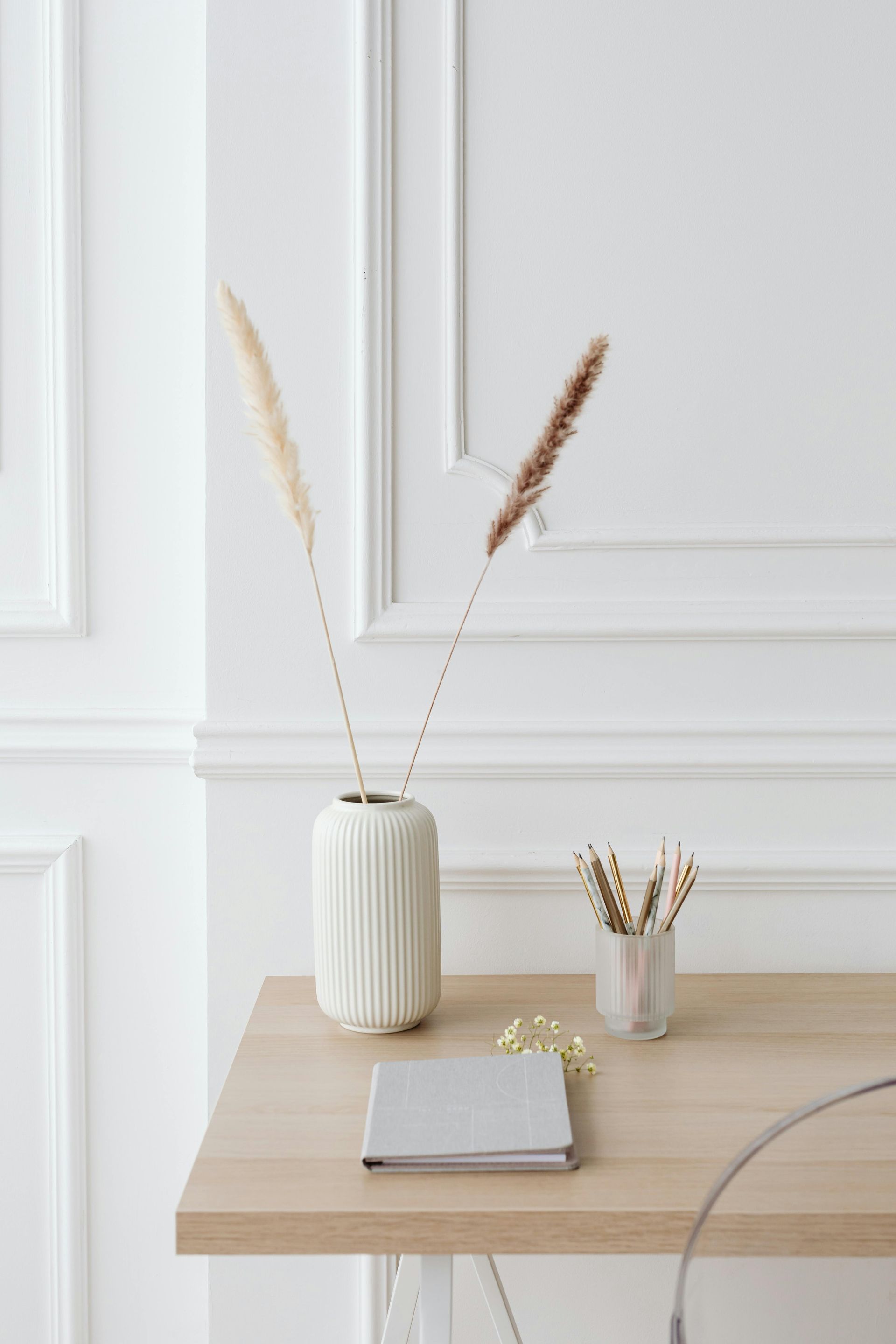 Desk with vase of pampas grass, small vase with dried stems, notebook. White wall with molding.