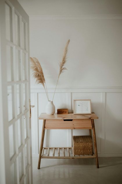 Wooden console table with vase of pampas grass, a framed picture, and a woven basket. Light-filled room.