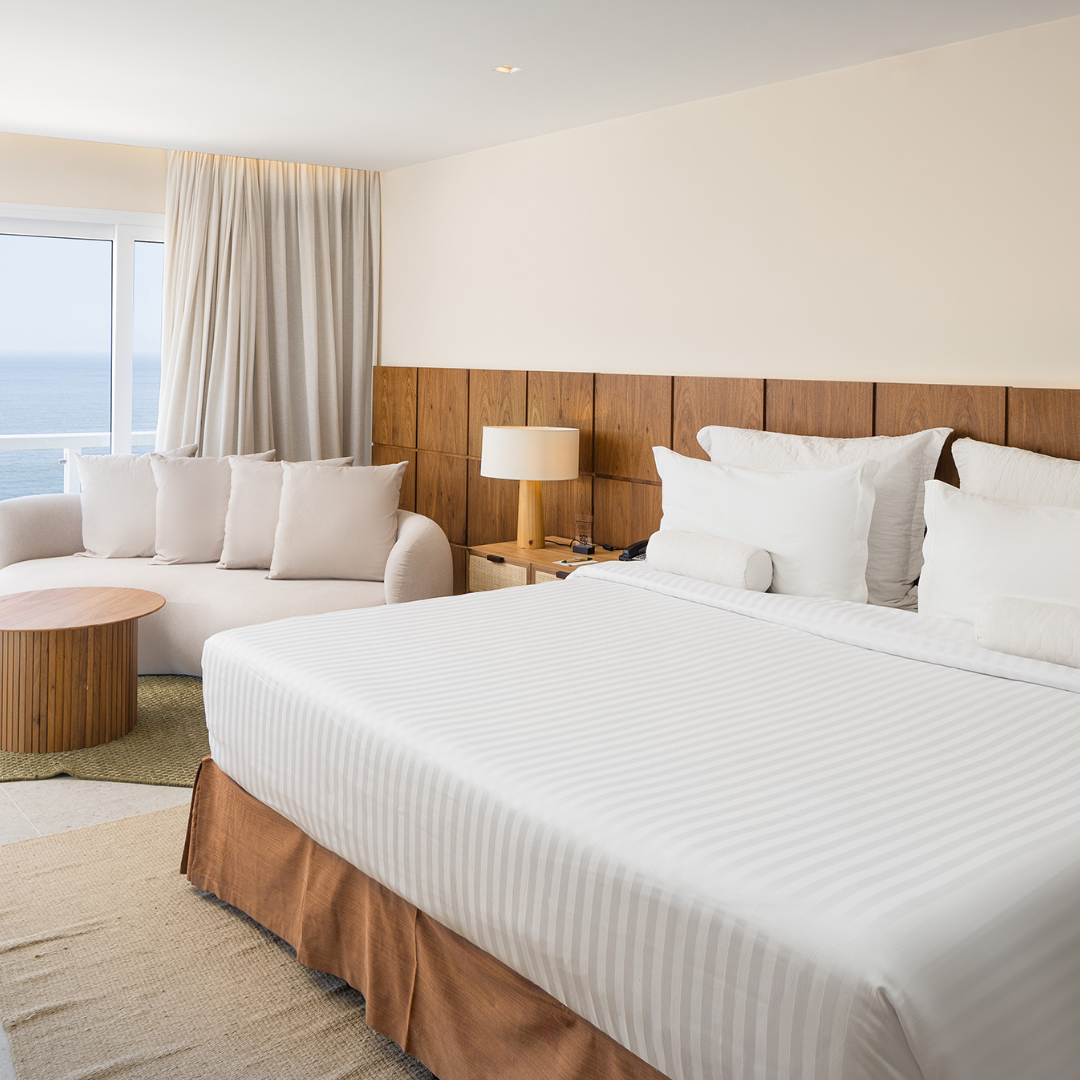 Quarto com cama, sofá, mesa de cabeceira e vista para o mar. Tons claros e neutros.