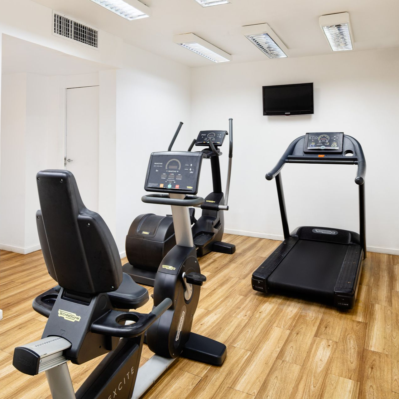 Academia com equipamentos de ginástica: bicicleta ergométrica, elíptico, esteira e TV.