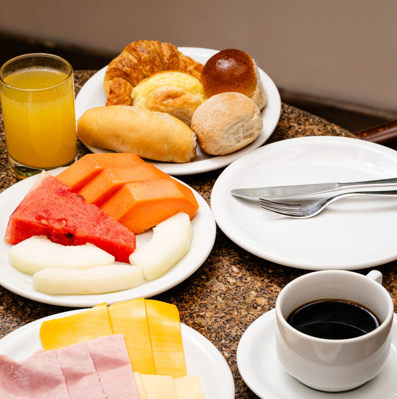 Café da manhã servido em uma mesa: suco, frutas, pão, presunto, queijo e café.