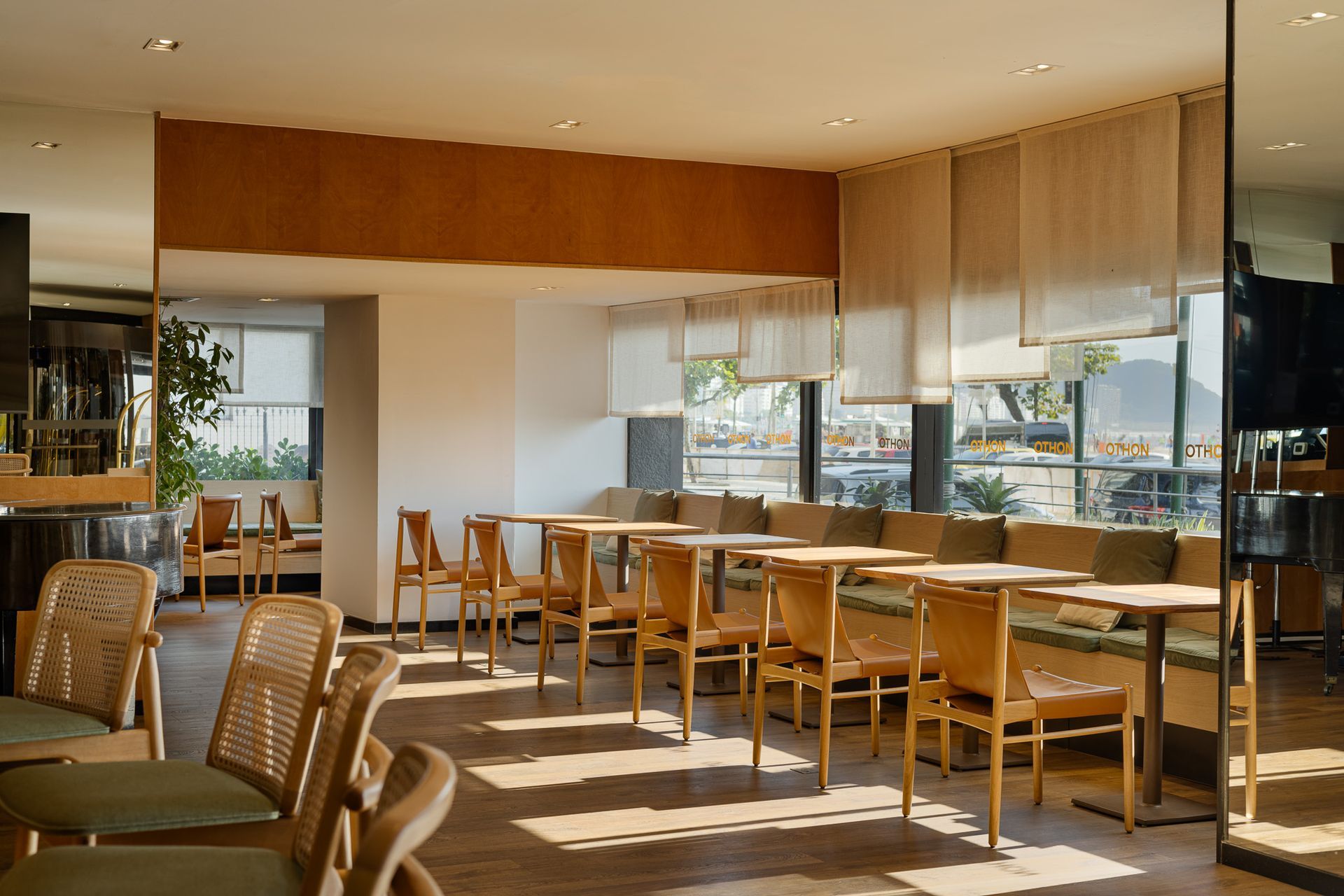 Interior de restaurante com mesas, cadeiras e grandes janelas. A luz do sol entra em abundância, iluminando o espaço.