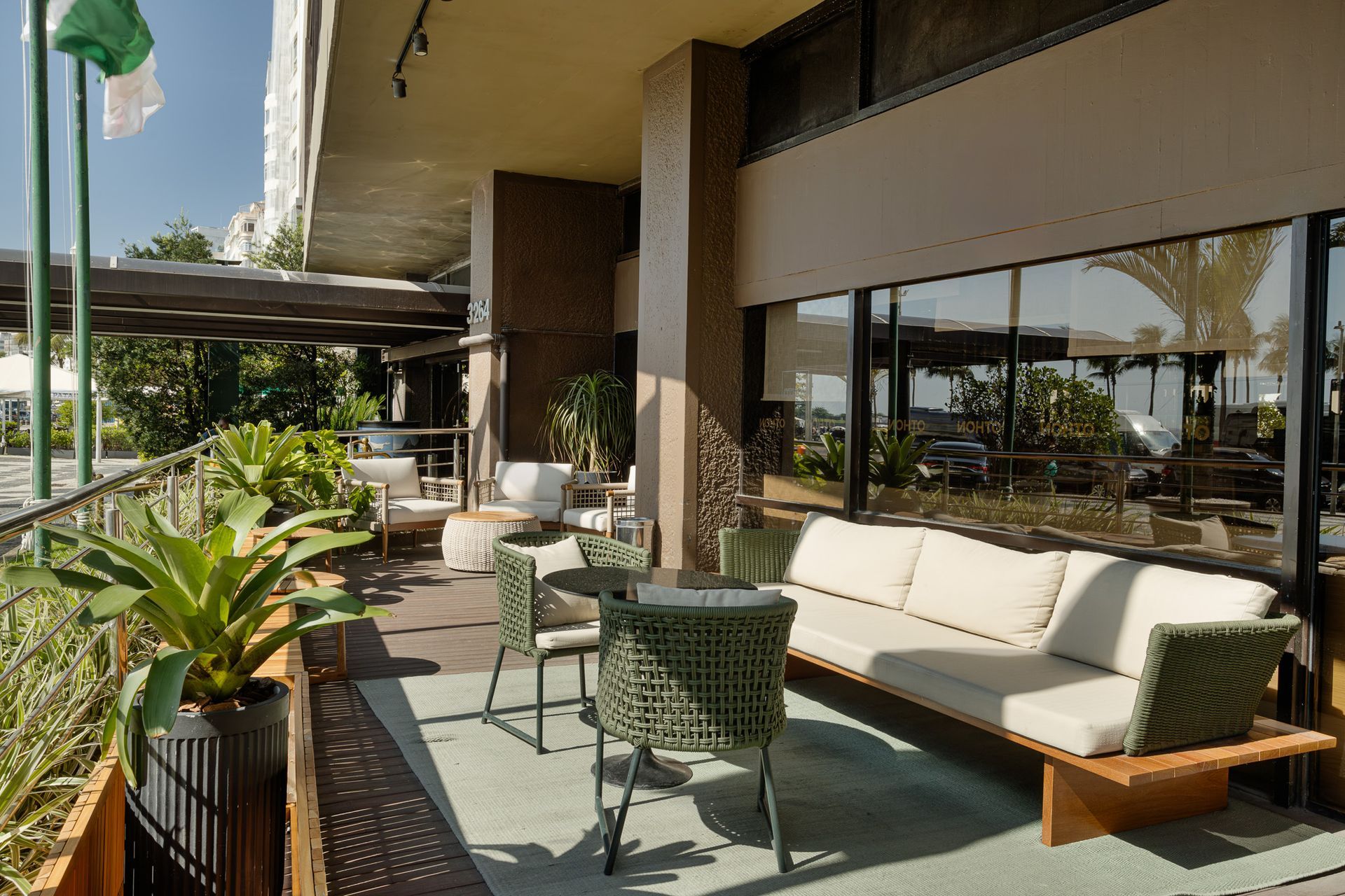 Pátio externo com área de estar: sofá, cadeiras, mesas e plantas, com vista através de grandes janelas.