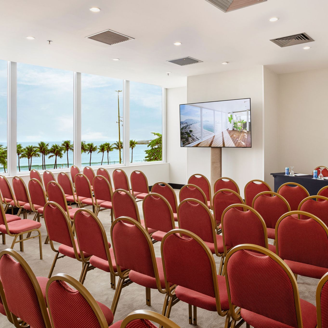 Fileiras de cadeiras vermelhas estão dispostas de frente para uma tela em uma sala de conferências com vista para o oceano.