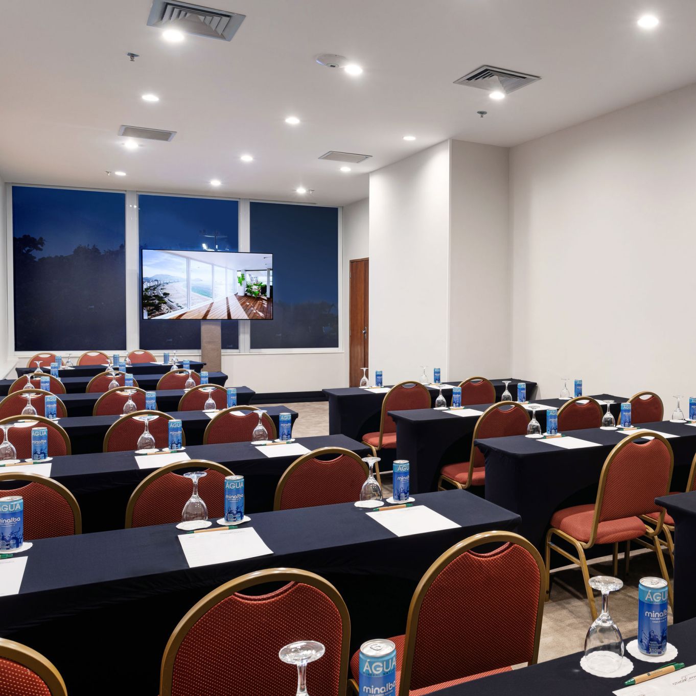 Sala de conferências com fileiras de mesas, cadeiras vermelhas e uma tela exibindo uma imagem.