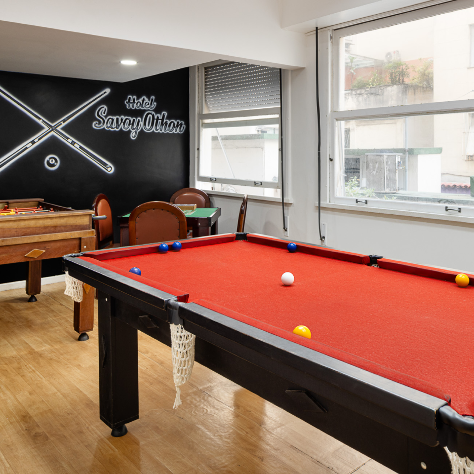 Sala de bilhar com mesa de bilhar de feltro vermelho, mesa de pebolim e uma parede preta com arte em neon de tacos de bilhar.