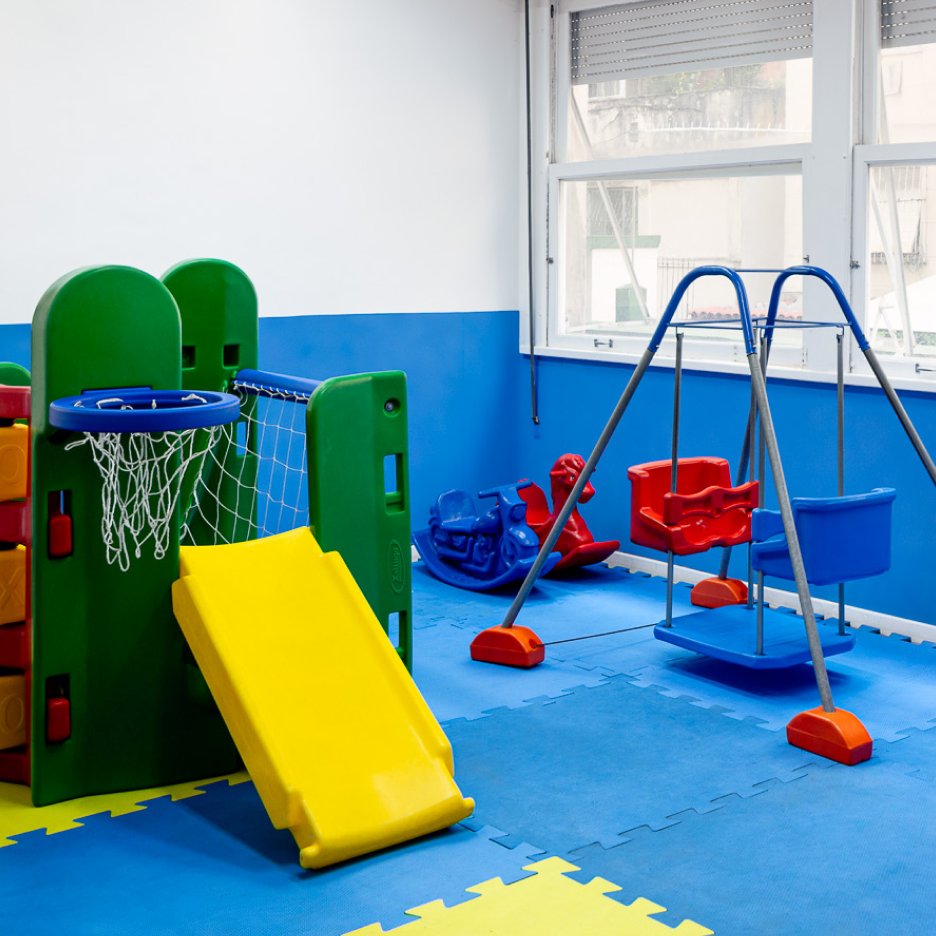 Sala de jogos com tapetes azuis e amarelos, balanços, escorregador, cesta de basquete e um cavalo de balanço.