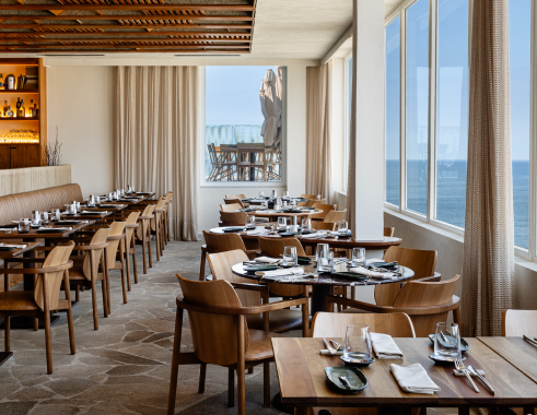 Interior do restaurante com mesas e cadeiras de madeira, grandes janelas com vista para o mar e paleta de cores neutras.