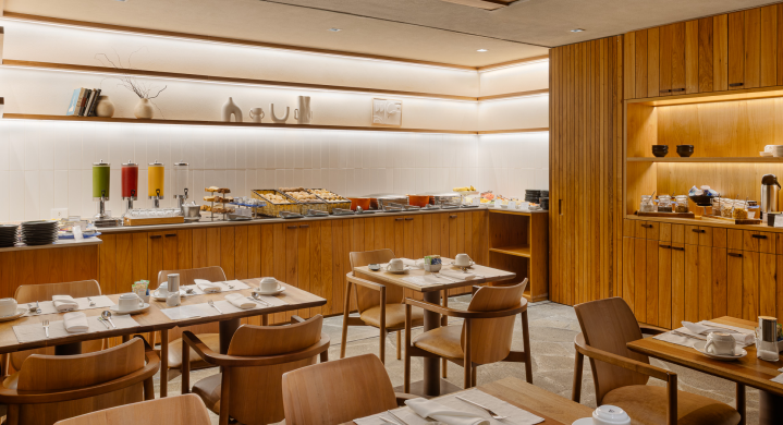 Área de café da manhã do hotel com balcões e mesas de madeira, buffet de comidas e cadeiras com lugares marcados.