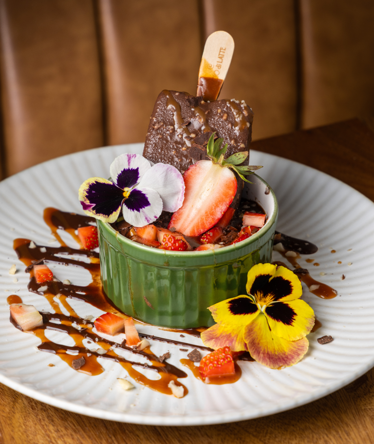 Sobremesa em ramequim verde: picolé de chocolate, morangos, flores comestíveis e calda de chocolate sobre prato branco.