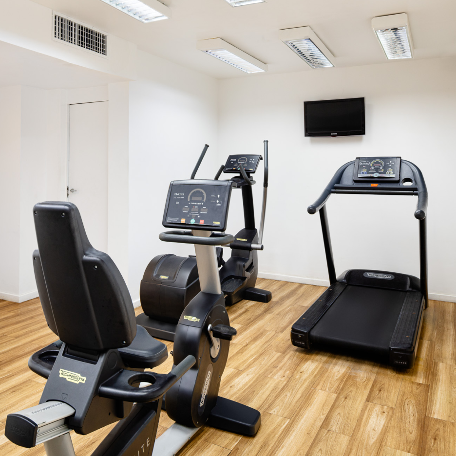 Academia com equipamentos de ginástica: bicicleta ergométrica reclinada, elíptico, bicicleta ergométrica vertical, esteira e TV em piso de madeira clara.