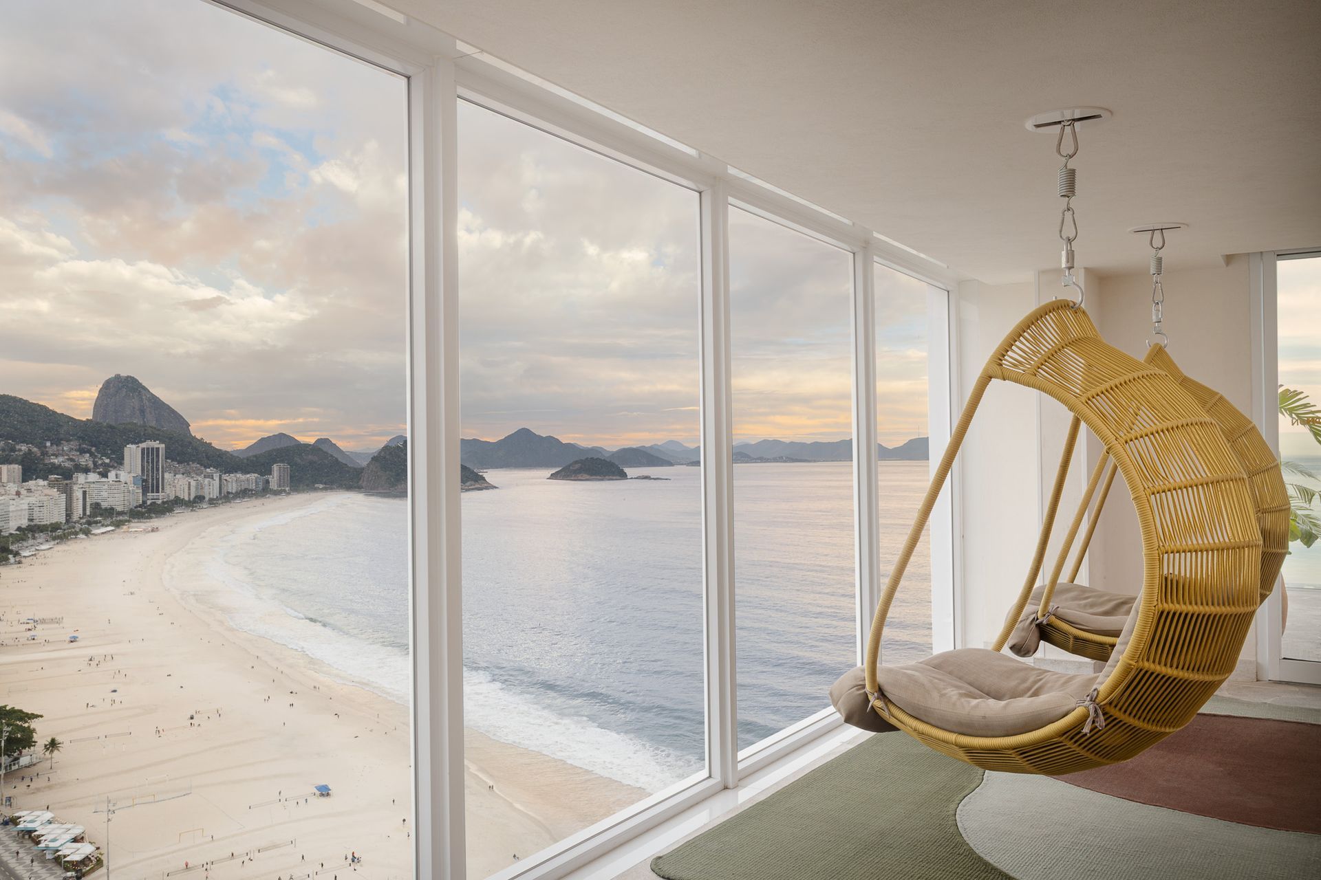 Uma cadeira de vime suspensa junto a uma grande janela com vista para a praia e o oceano de Copacabana.