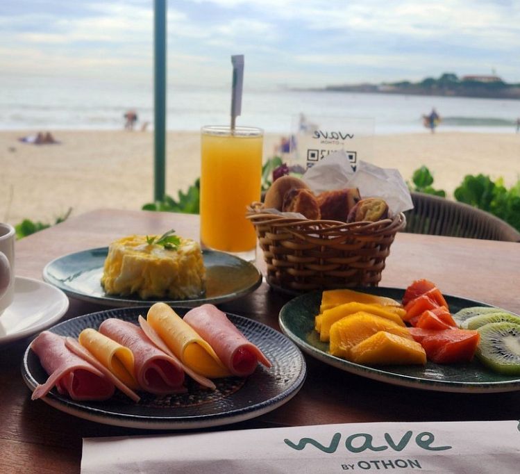 Café da manhã servido em uma mesa com vista para a praia: ovos, presunto, frutas, suco, cesta de pães e um código QR.