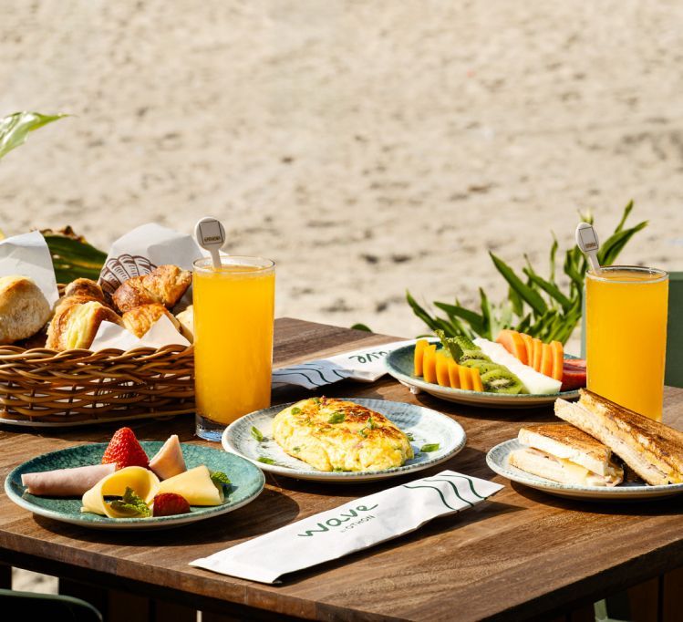 Café da manhã servido em uma mesa ao ar livre, com frutas, ovos, torradas, suco e pão, tendo como pano de fundo a areia da praia.