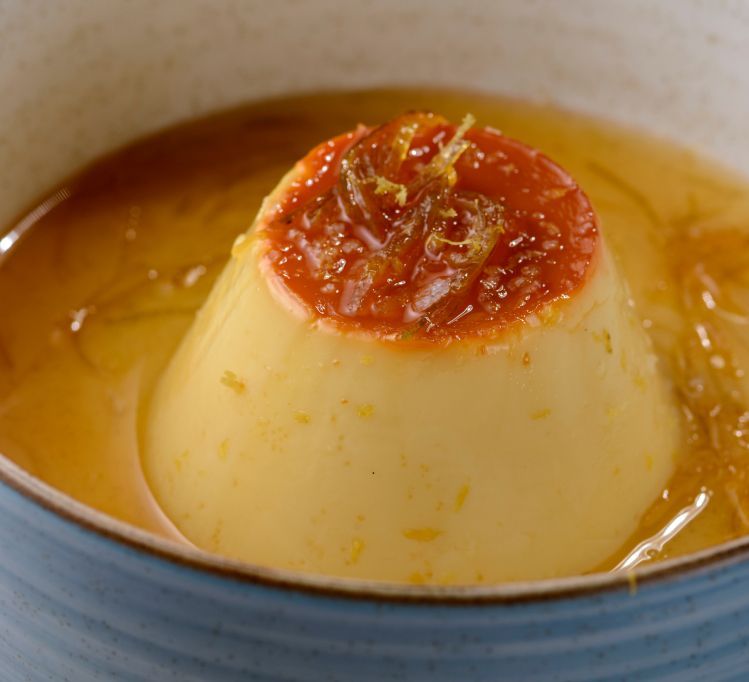 Sobremesa de creme de baunilha em calda de laranja, decorada com casca de laranja cristalizada, servida em uma tigela azul.