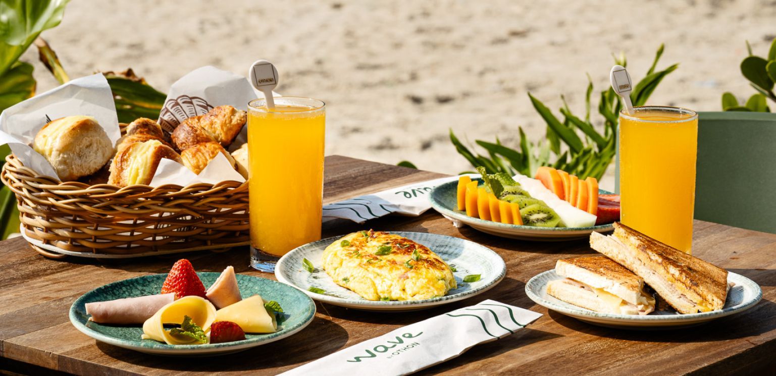 Café da manhã servido em uma mesa de praia: cesta de pães, pratos de frutas, omelete, copos de suco e sanduíches tostados.