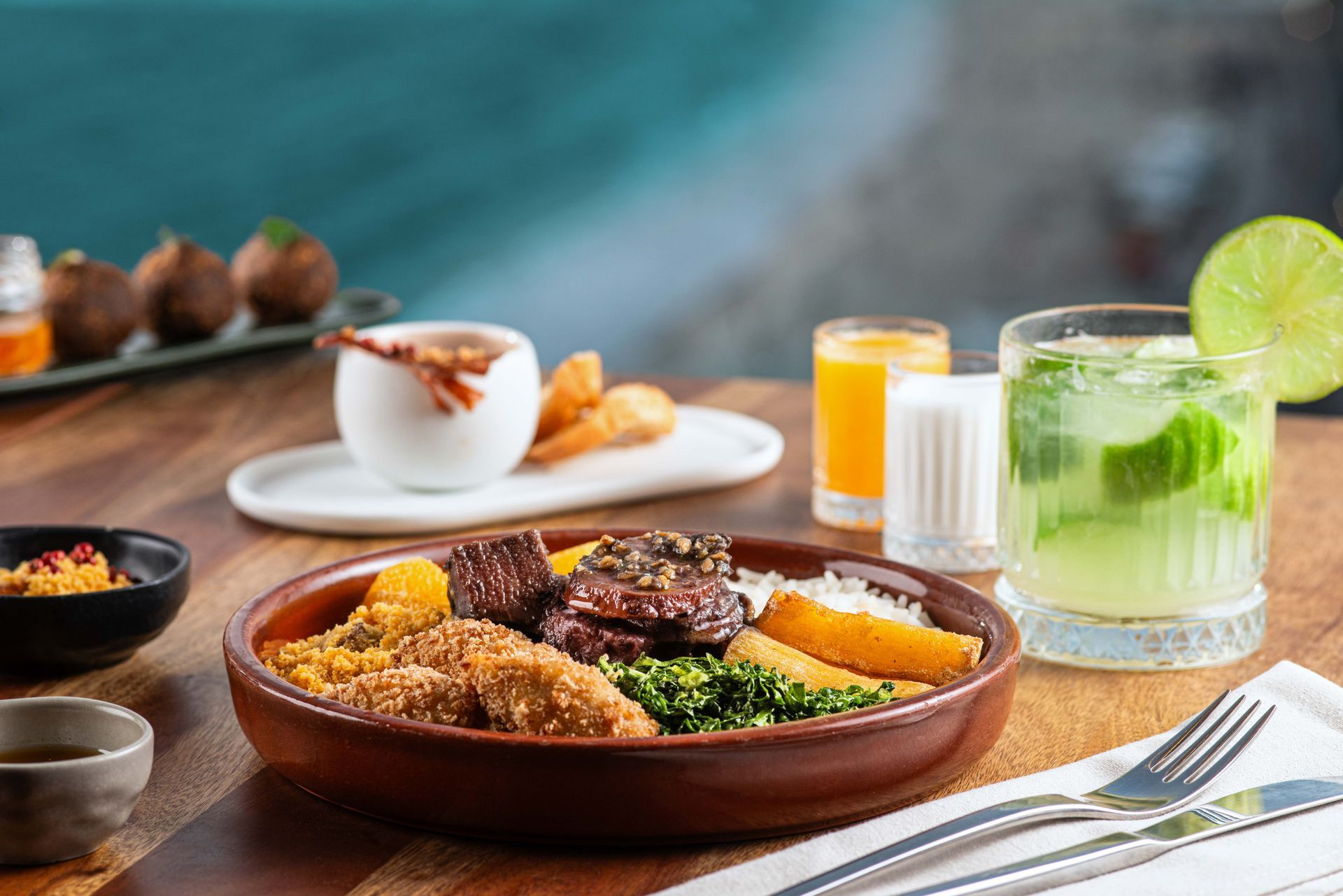 Refeição brasileira: carne, arroz, verduras, batatas fritas, caipirinha, suco de laranja, aperitivos e condimentos sobre uma mesa de madeira.