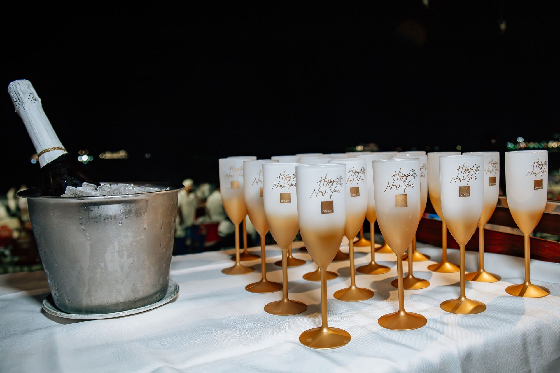 Taças de champanhe enfileiradas sobre uma mesa, ao lado de um balde de gelo com uma garrafa. Fundo escuro. Othon