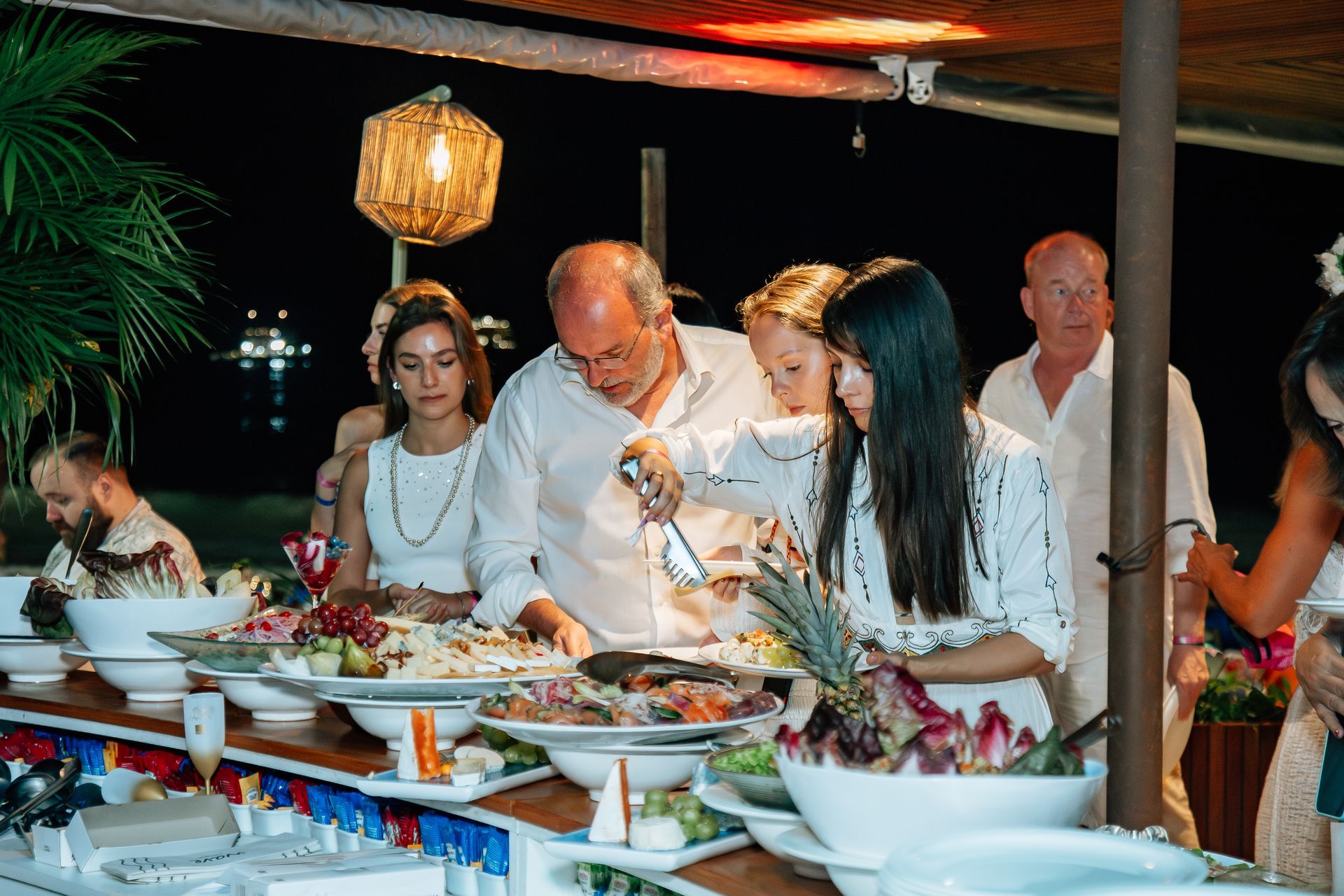 Pessoas em uma mesa de bufê, escolhendo comida. Mesa posta ao ar livre à noite. Muitos pratos, roupas brancas. Othon
