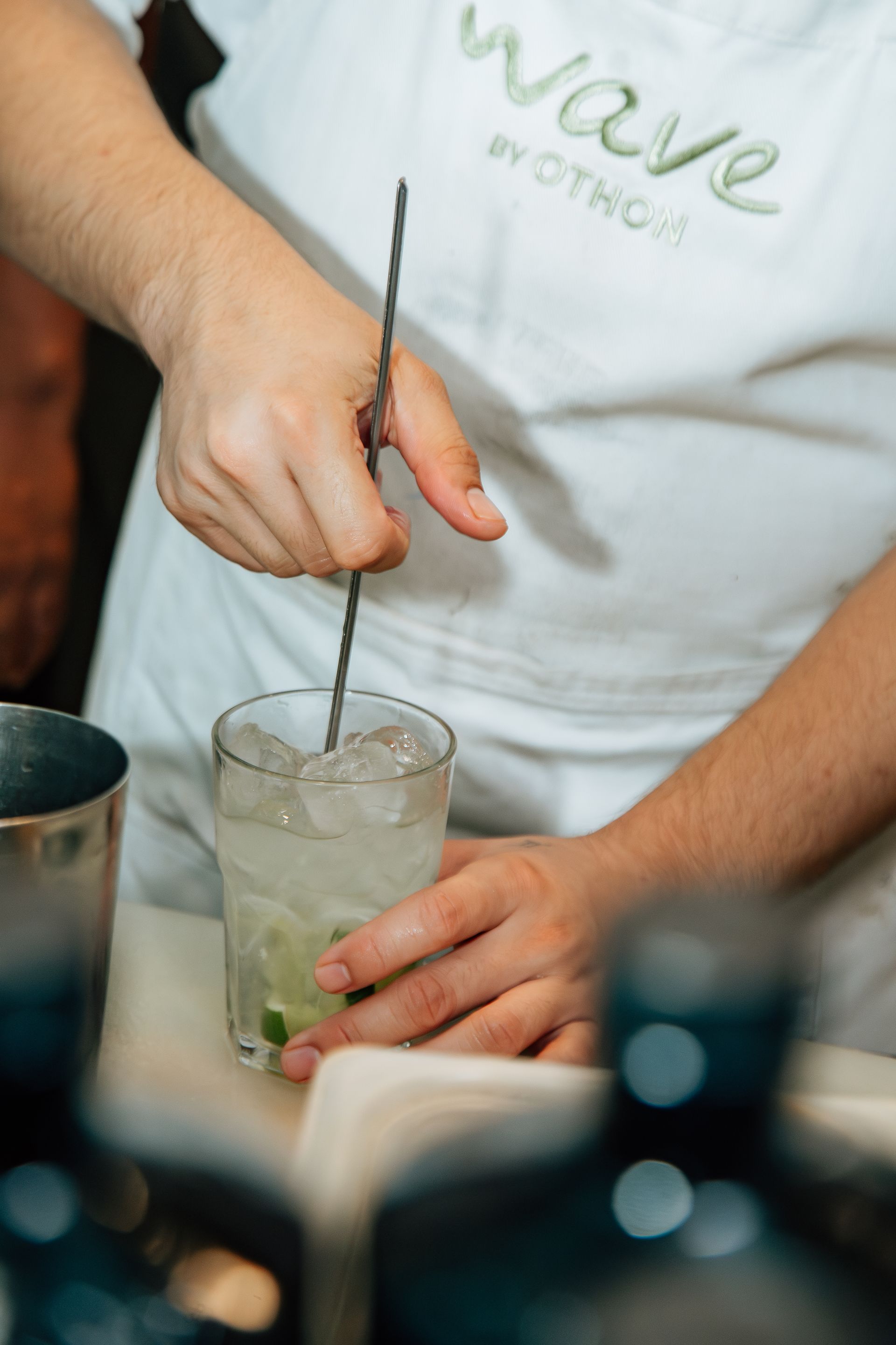 Barman mexendo coquetel com gelo e limão, usando avental branco com os dizeres 
