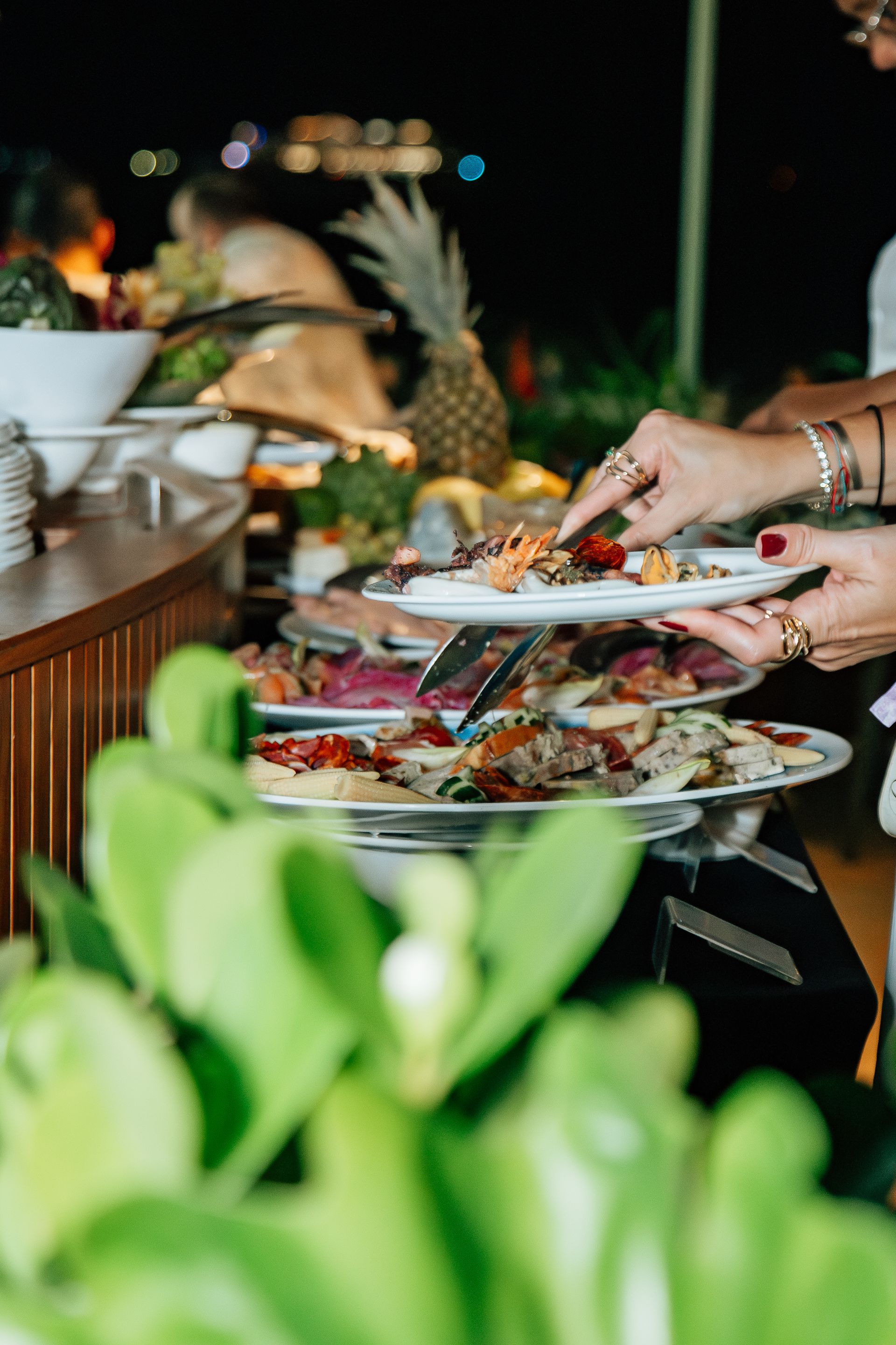Buffet com diversos pratos de comida. Uma pessoa usa pinças para se servir. Folhagem verde em primeiro plano. Othon