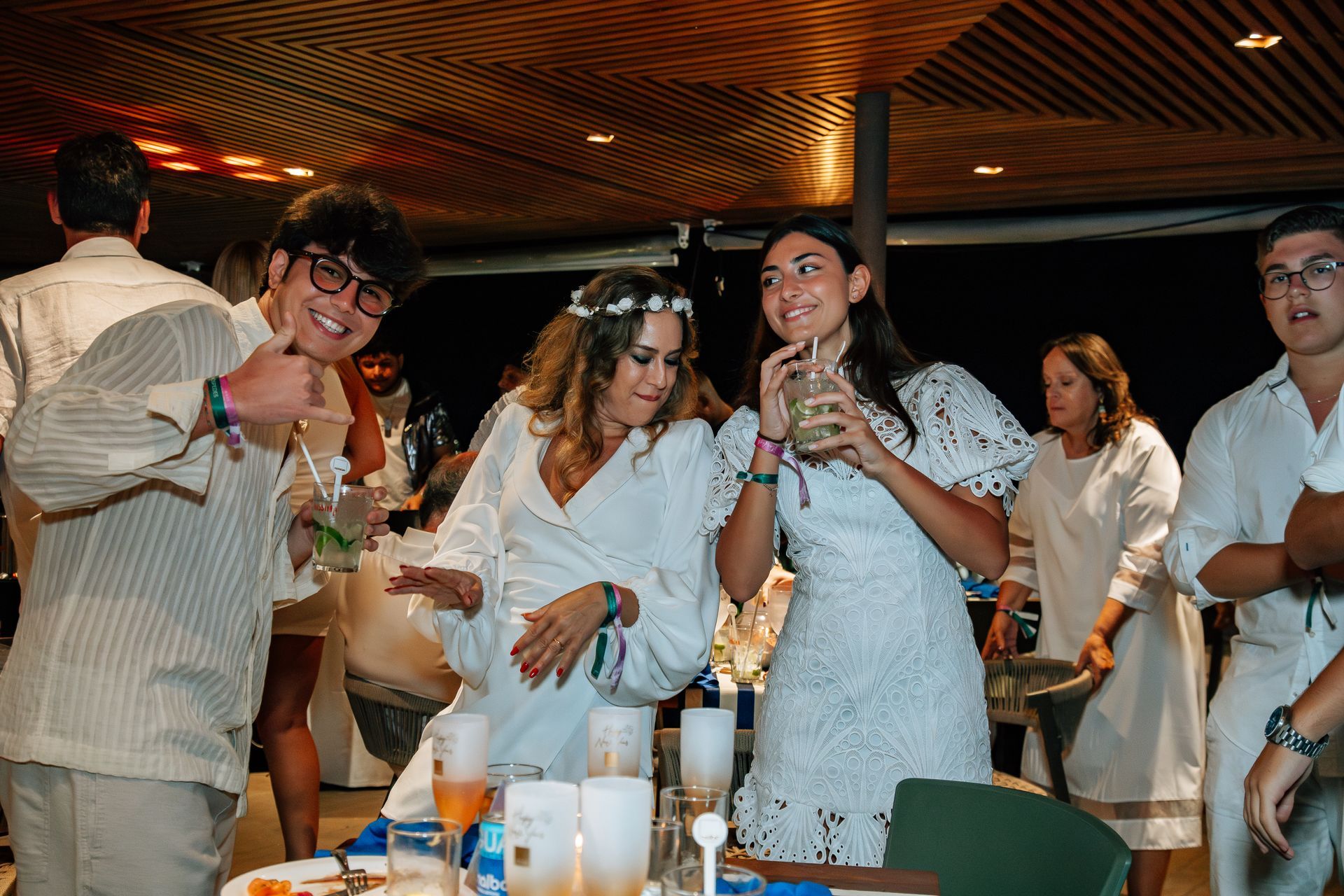 Grupo de pessoas dançando em uma festa noturna, todas vestidas de branco. Algumas seguram bebidas e sorriem. Othon