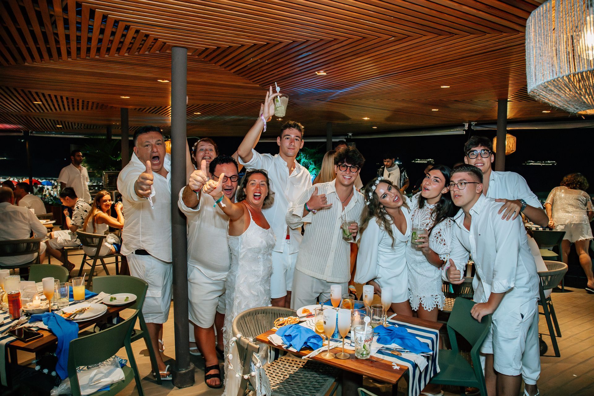 Grupo de pessoas vestidas de branco comemorando em uma mesa em um restaurante com teto de madeira e detalhes em azul. Othon