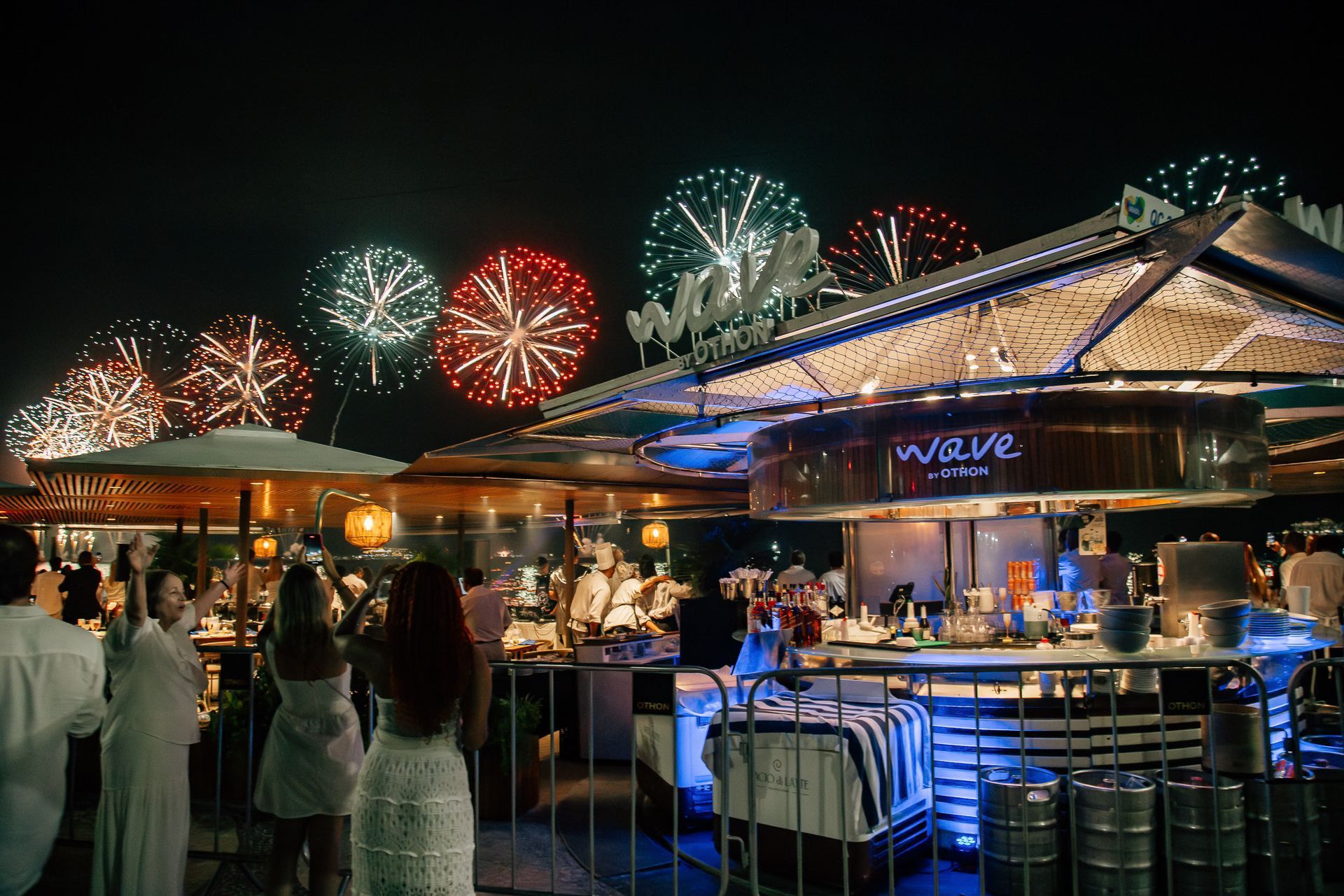 Fogos de artifício sobre um restaurante com clientes, bar iluminado e placa 