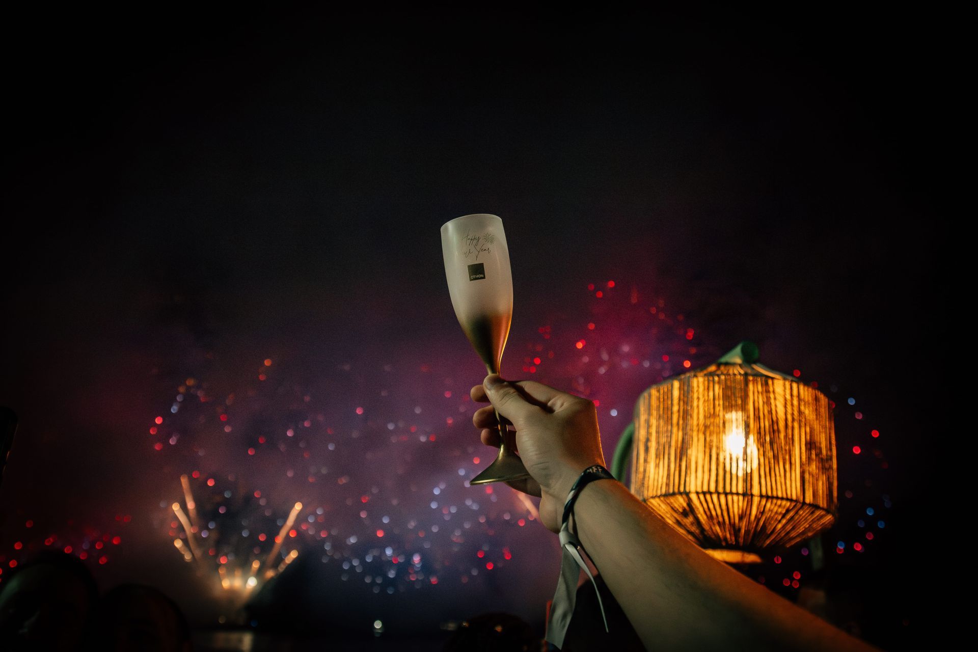 Mão segurando uma taça de champanhe, fogos de artifício ao fundo. Lanterna com luz quente à direita. Othon