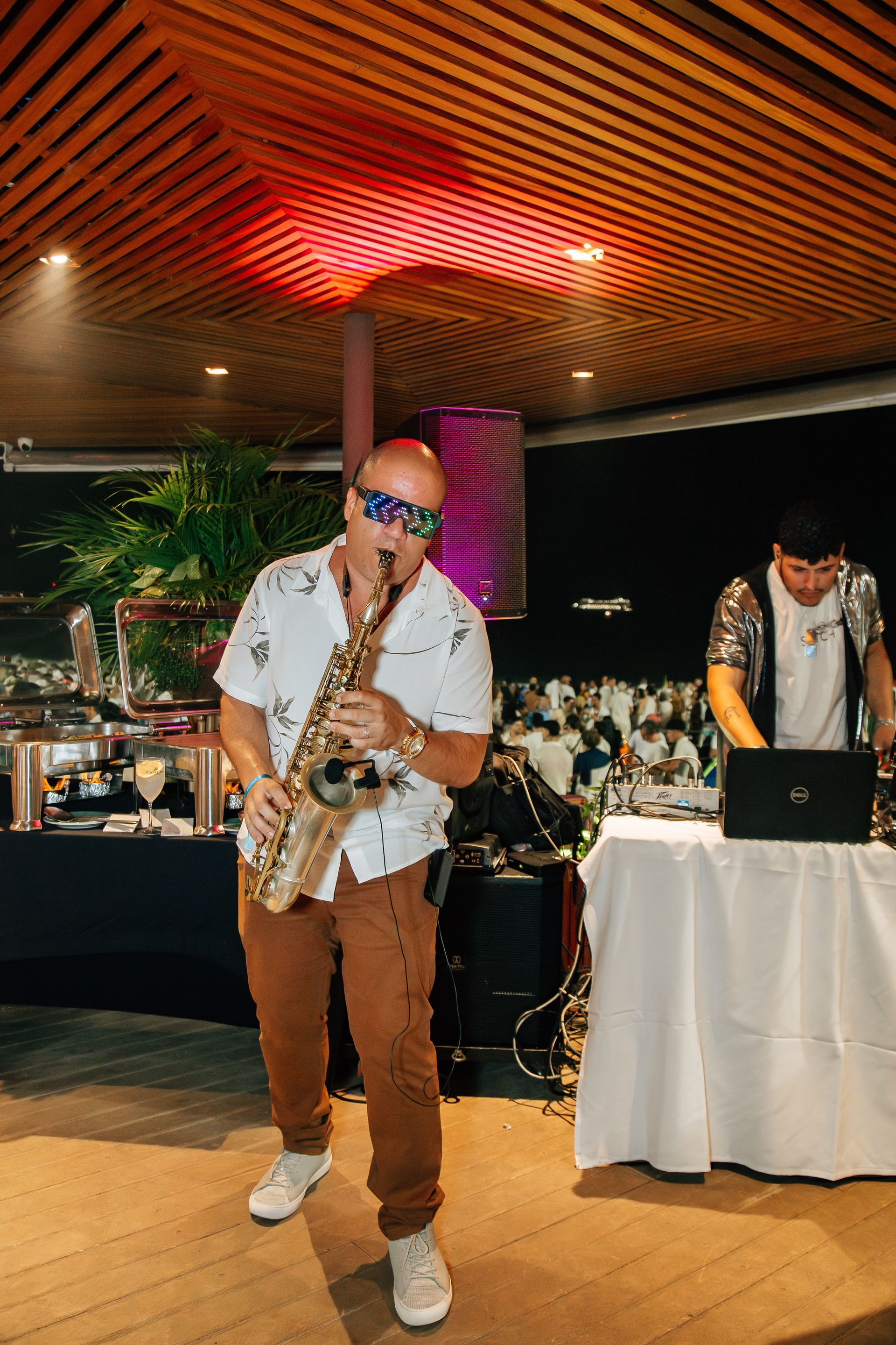 Homem careca de óculos escuros toca saxofone e é DJ atrás de uma mesa em um local mal iluminado. Othon