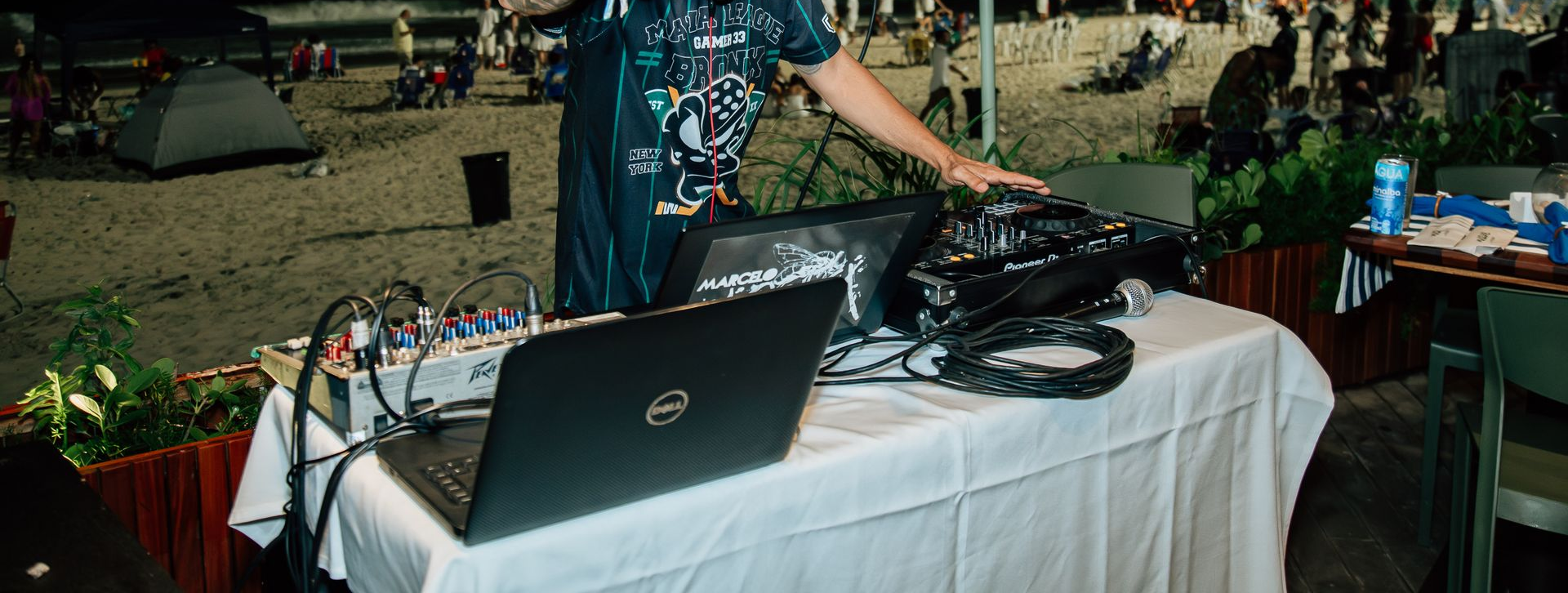 Um DJ fica atrás de equipamentos em um evento ao ar livre, com um laptop, um mixer e outros equipamentos em uma mesa coberta de branco. Othon