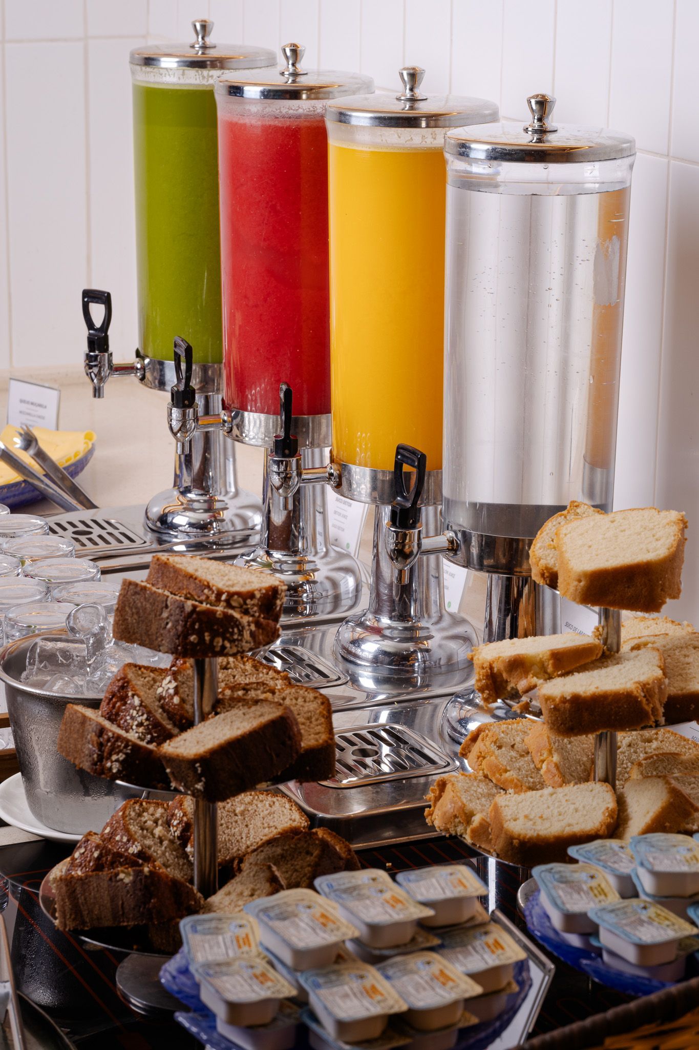 Dispensadores de suco com líquidos verdes, vermelhos, amarelos e transparentes; diversos tipos de pães; e pacotes de manteiga.