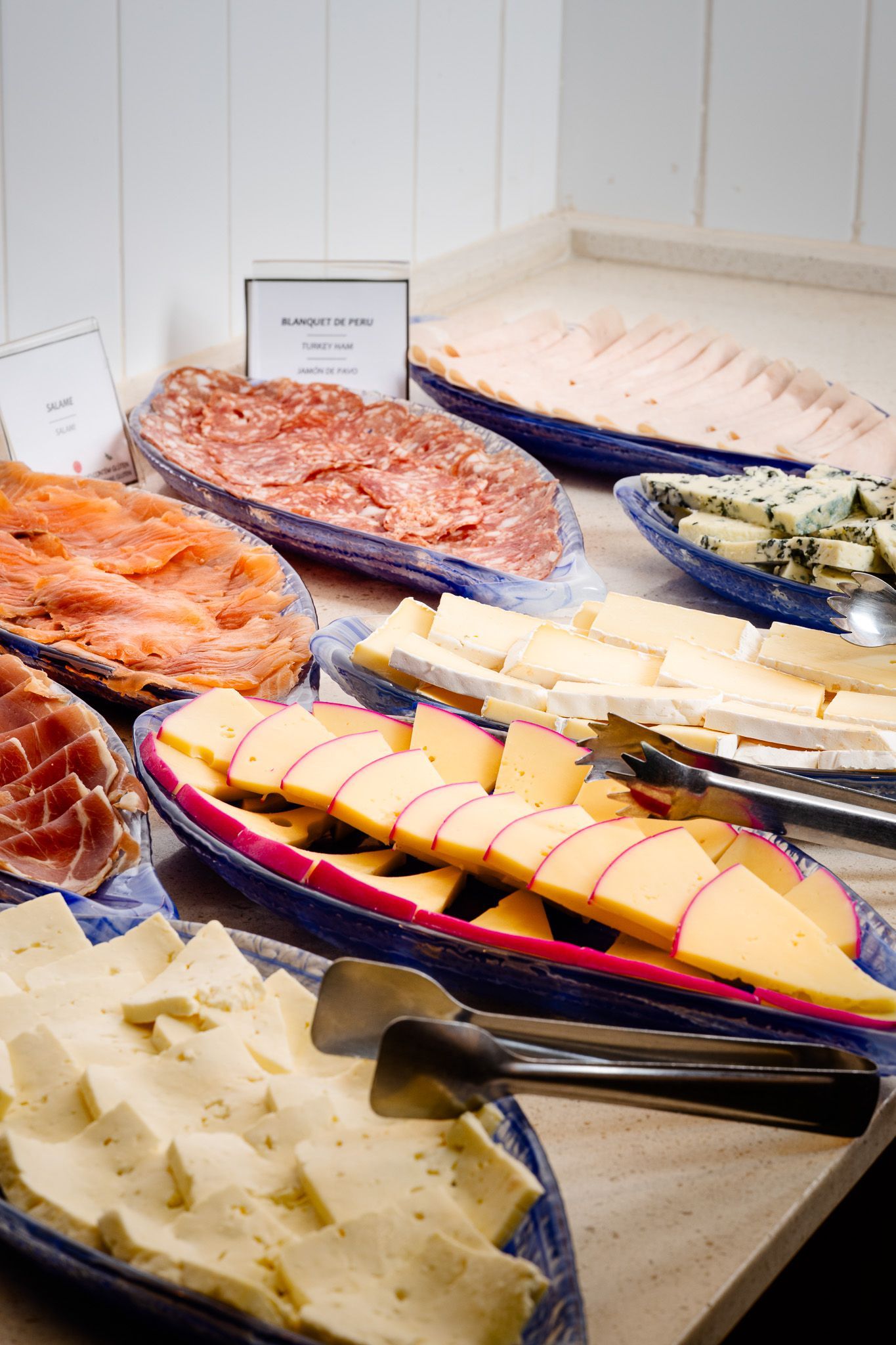Buffet com diversos queijos, carnes e salmão defumado em travessas azuis, iluminado por cima.