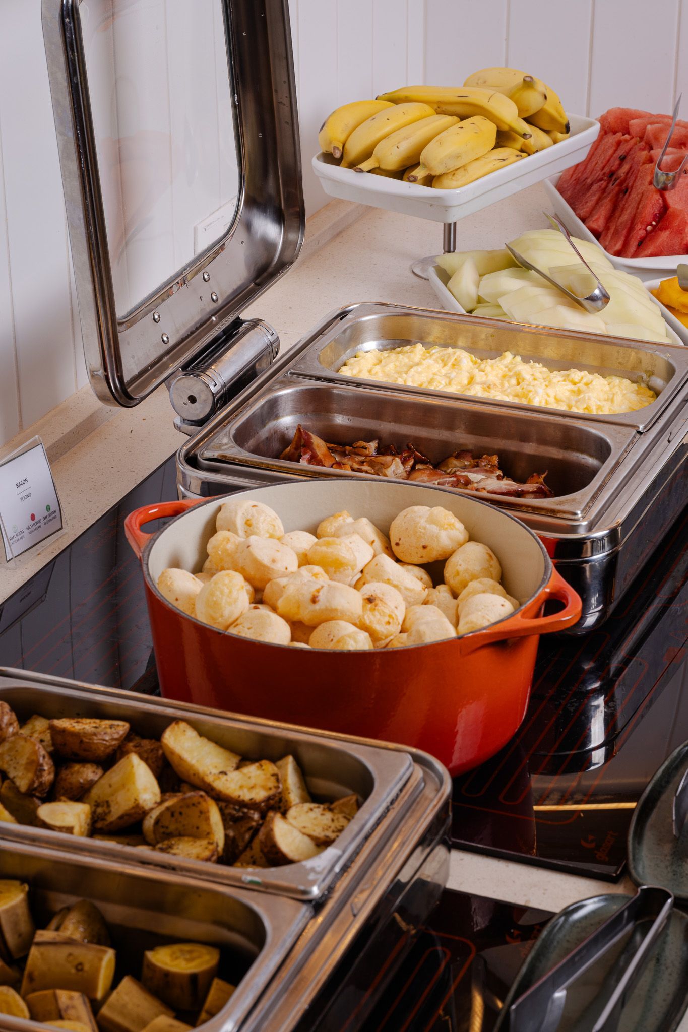 Buffet de café da manhã com diversos itens: batatas, pão com queijo, bacon, ovos mexidos e frutas.