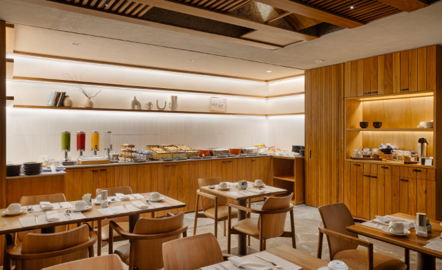 Área de buffet de café da manhã do hotel com móveis de madeira, alimentos e mesas postas para refeições.