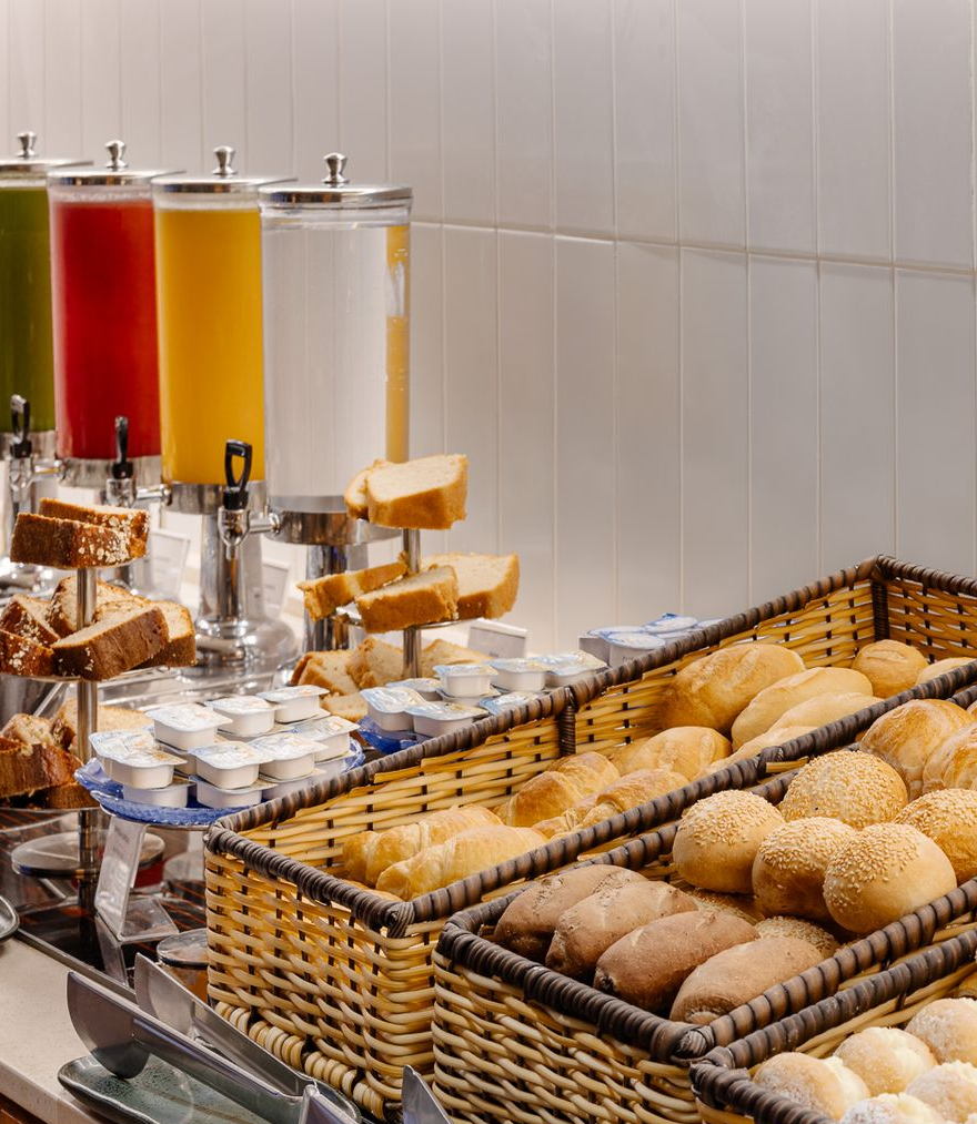 Buffet de café da manhã com máquinas de suco, pães e pãezinhos.