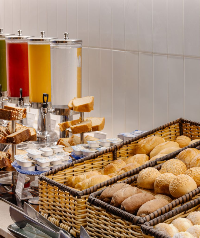 Buffet de café da manhã com máquinas de suco, pães e pãezinhos.