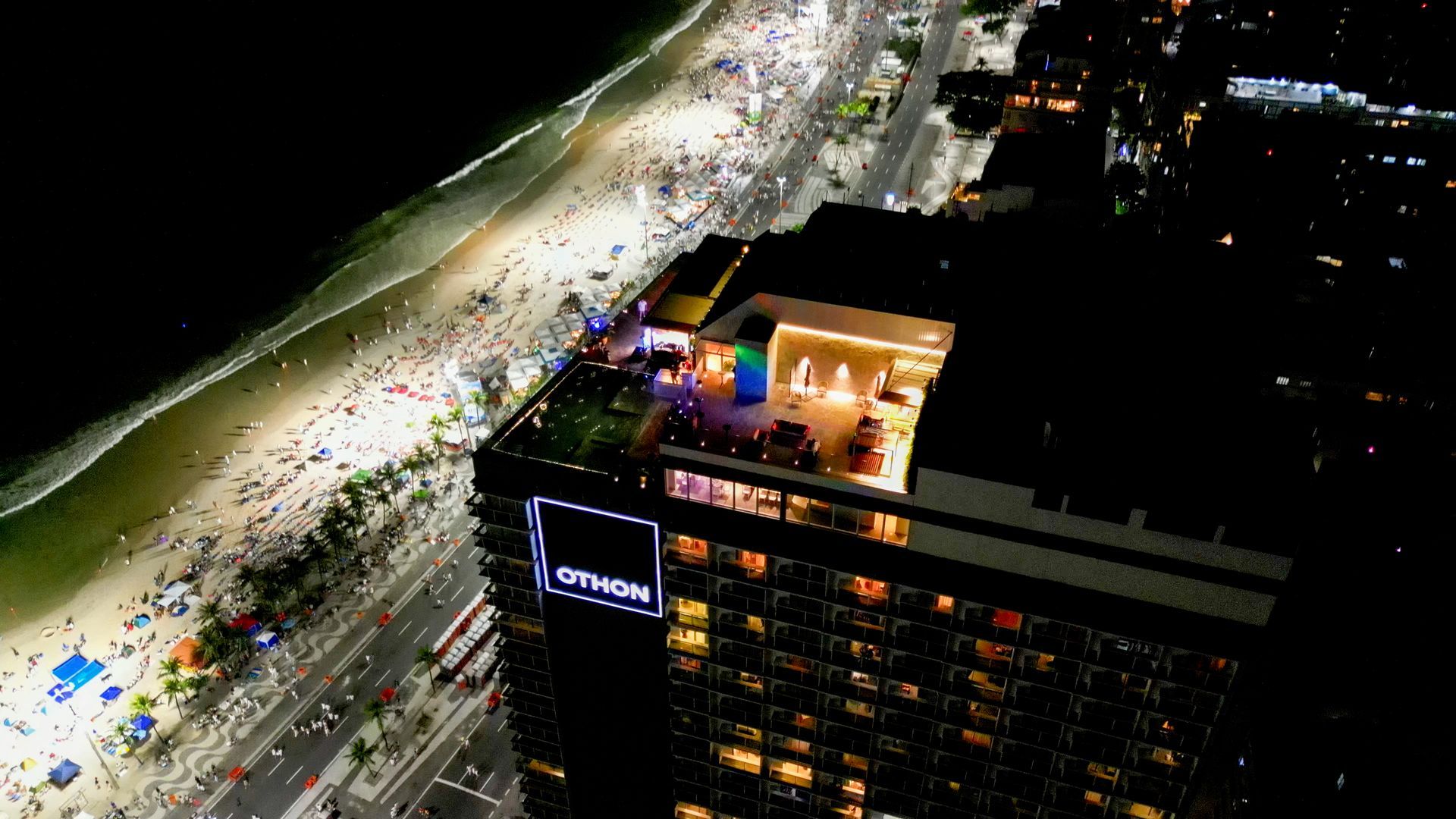 Vista aérea noturna de um prédio alto com um bar na cobertura com vista para uma praia movimentada e para o oceano. Othon