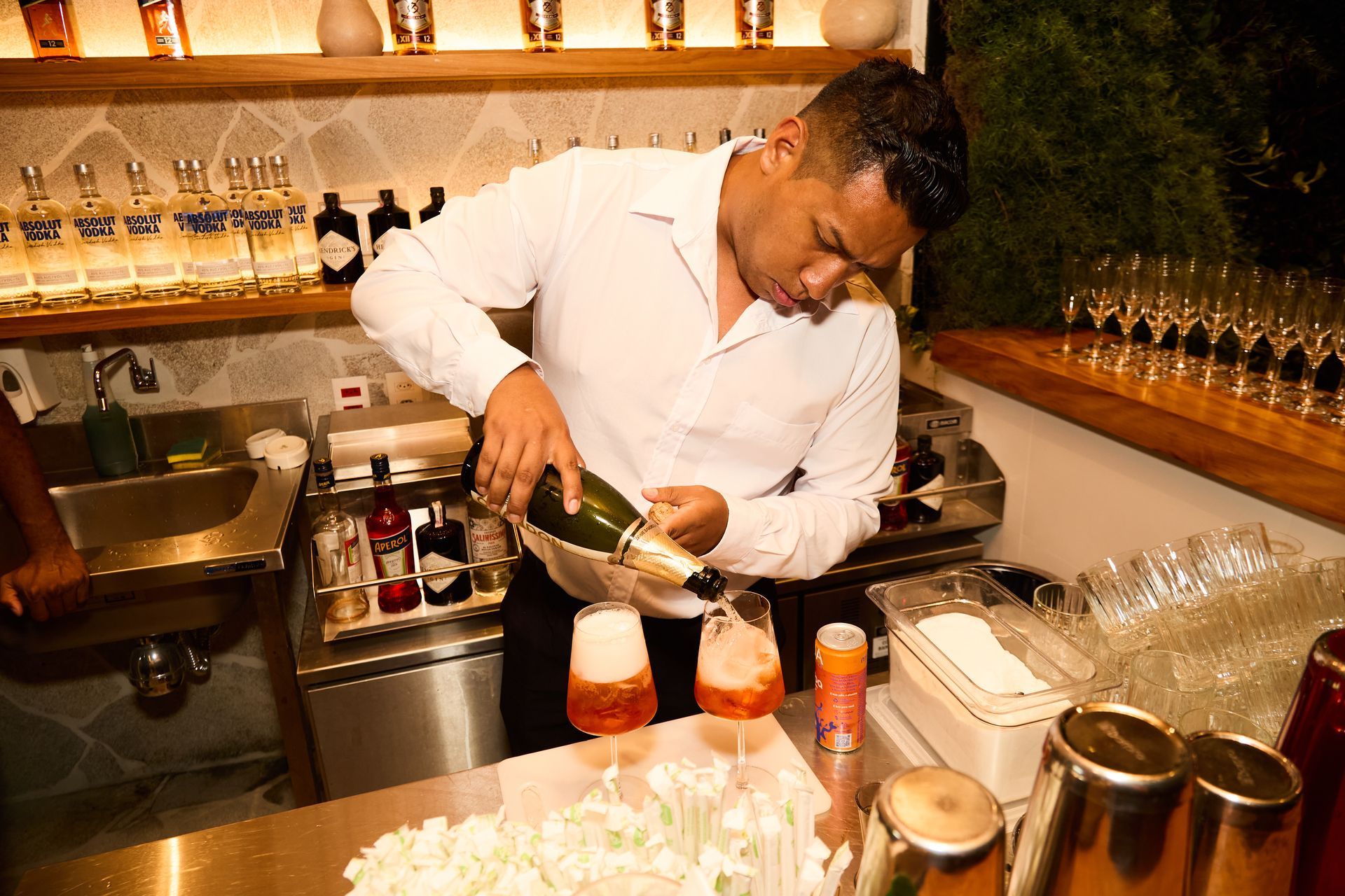 Barman servindo champanhe em taças, preparando um coquetel. Ambiente de bar com garrafas e taças. Othon