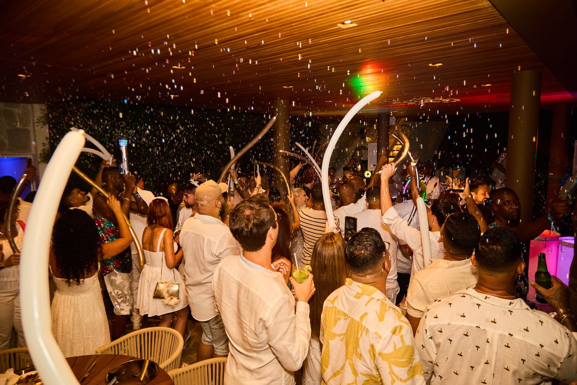 Cena de festa com confete caindo sobre uma multidão vestida de branco, iluminada por luzes coloridas. Othon